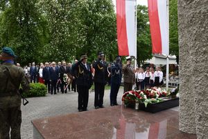 Zdjęcie w kolorze przedstawia, delegację złożoną z komendantów powiatowych Policji , Straży Pożarnej oraz Nadleśnictwa w Oleśnie. Którzy oddają honor przy pomniku. W tle pozostałe delegacje, zgromadzeni mieszkańcy. Po prawej stronie biało czerwone flagi.