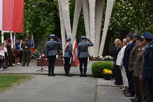 Zdjęcie w kolorze przedstawia, poczet sztandarowy Komendy Powiatowej Policji w Oleśnie podczas oddania honoru w stronę pomnika, po lewej stronie biało czerwone flagi, po lewej mieszkańcy i delegacje.