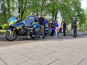 Zdjęcie w kolorze przedstawia na pierwszym planie motocykl policyjny, za którym stoją dorośli oraz dzieci w towarzystwie policjantów. Tuz za nimi radiowóz policyjny i plakat rozwijany na stojaku z napisem: rekrutacja trwa , dołącz do nas. W tle drzewa.