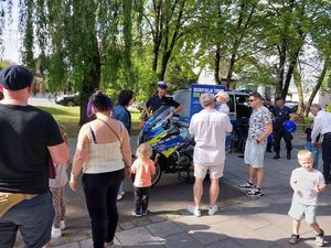 Zdjęcie w kolorze przedstawia dorosłych oraz dzieci w towarzystwie policjantów. Po lewej stronie policyjny motocykl. Z tyłu radiowóz policyjny i plakat rozwijany na stojaku z napisem: rekrutacja trwa , dołącz do nas. W tle drzewa.