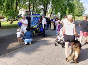 Zdjęcie w kolorze przedstawia na pierwszym planie  osoby przechodzące z psem po lewej stronie motocykl policyjny, za którym stoją dorośli oraz dzieci w towarzystwie policjantów. Tuz za nimi radiowóz policyjny, W tle drzewa.