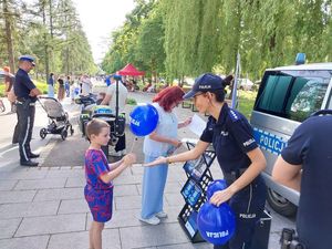 Zdjęcie w kolorze przedstawia na pierwszym planie policjantka wręczająca młodemu chłopcu niebieski balon z napisem policja. W tle uczestnicy pikniku przebywający na promenadzie.
