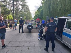 Zdjęcie w kolorze przedstawia na pierwszym planie po prawej policjantów stojących przy radiowozie, na wprost policyjny motocykl, na którym siedzi mała dziewczynka w asyście policjanta. W tle uczestnicy pikniku przebywający na promenadzie.
