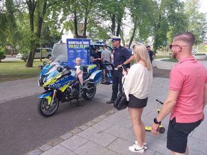 Zdjęcie w kolorze przedstawia na pierwszym planie dorosłych mężczyznę i kobietę dalej po lewej motocykl policyjny, na którym siedzi mały chłopiec w towarzystwie policjanta.  Tuz za nimi radiowóz policyjny i plakat rozwijany na stojaku z napisem: rekrutacja trwa , dołącz do nas. W tle drzewa.