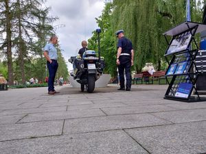 Zdjęcie w kolorze przedstawia na pierwszym planie po prawej półka z materiałami promocyjnymi. dalej na środku policjant motocykl policyjny, na którym siedzi mały chłopiec w towarzystwie policjanta po prawej i mężczyzny po lewej. W tle uczestnicy pikniku przebywający na promenadzie.