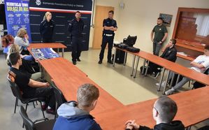zdjęcie w kolorze przedstawia uczniów siedzących przy stole ułozonym w okrąg w sali narad KPP w Oleśnie, na środku stoją policjanci, Za nimi płócienna ścianka na stelażu z napisem policja olesno.