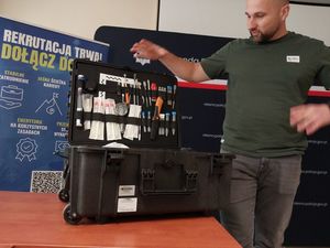 Zdjęcie w kolorze przedstawia mężczyznę ubranego po cywilnemu, który omawia zawartosc leżącej obok niego walizki technika kryminalistyki, w tle plakat rozwijany z roli z napisem &quot;dołącz do nas rekrutacja trwa&quot;.