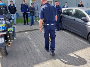 Zdjęcie w kolorze przedstawia uczniów stojących na placu zewnętrznym KPP w Oleśnie, na środku policjant demonstruje urzadzenie do pomiaru odległosci, po lewej stronie stoi motocykl.