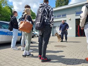 Zdjęcie w kolorze, przedstawia uczniów na placu zewnętrznym KPP w Olesnie, na środku policjant z psem. W tle garaż.