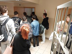 Zdjęcie w kolorze przedstawia uczniów wewnątrz pomieszczenia dla osób zatrzymanych, z prawej strony krata zabezpieczająca drzwi wejściowe.