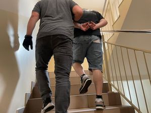 zdjęcie w kolorze przedstawia dwóch mężczyzn idących schodami w górę, mężczyzna po lewej stronie to nieumundurowany policjant, który trzyma pod rękę drugiego mężczyznę, który ma kajdanki założone na ręce trzymane z tyłu.