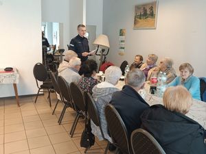 Zdjęcie w kolorze. przedstawia osoby siedzące przy podłużnym stole w pomieszczeniu. Na pierwszym planie po prawej stronie siedzą seniorzy. Na końcu stołu po prawej stoi policjant.