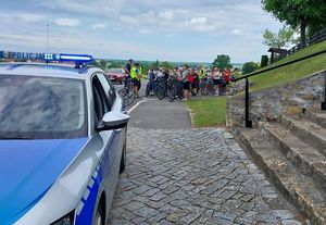 Zdjęcie w kolorze przedstawia na pierwszym planie po lewej stronie fragment radiowozu, za nim peleton rowerzystów zatrzymujących się na parkingu przy kościele pw. Św. Roha. W tle panorama olesna oraz niebo.