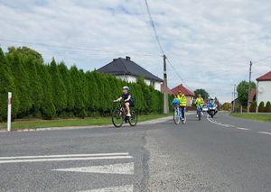 Zdjęcie w kolorze przedstawia rowerzystów zjeżdżających na skrzyżowaniu , tuż za nimi motocykl policyjny z włączonymi światłami błyskowymi.