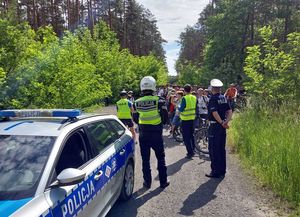 Zdjęcie w kolorze przedstawiana piwreszym planie po lewej fragment radiowozu dalej za nim policjanci i dojeżdżający leśną droga rowerzyści. w tle las.