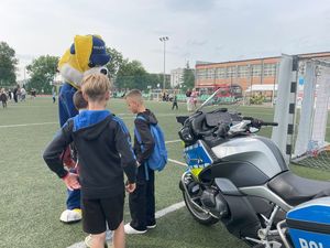 Zdjęcie w kolorze, przedstawia młodych chłopców stojących przy maskotce &quot;policusia&quot; , obok nich po prawej zaparkowany policyjny motocykl. W tle pozostali uczestnicy festynu.