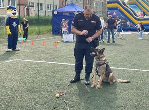 Zdjęcie w kolorze przedstawia policjanta - przewodnika wraz ze swoim psem, po lewej stronie stoi policus a w tle inne atrakcje dla dzieci, miedzy innymi &quot;dmuchańce&quot;.