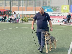 Zdjęcie w kolorze przedstawia policjanta przewodnika, który idzie w stronę obiektywu ze swoim psem.