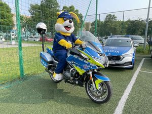 Zdjęcie w kolorze, przedstawia maskotkę &quot;policusia&quot; który siedzi na policyjnym motocyklu. Obok po prawej stronie zaparkowany radiowóz.
