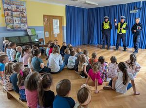 Zdjęcie w kolorze, przedstawia grupę kilkudziesięciu przedszkolaków siedzących na parkiecie, przed nimi stoi troje funkcjonariuszy ( dwóch mężczyzn i kobieta ). za nimi na ścianie błekitna zasłona.