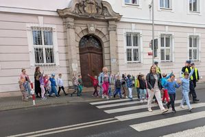 Zdjęcie w kolorze, przedstawia przedszkolaków przechodzących po przejściu dla pieszych w asyście opiekunek i policjantów. W tle fragment  budynku Sądu Rejonowego w Olesnie.