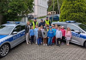 Zdjęcie w kolorze przedstawia grupę przedszkolaków pozujących do zdjęcia stojąc pomiędzy dwoma radiowozami. Za przedszkolakami stoją policjanci.