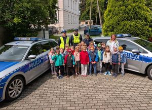 Zdjęcie w kolorze przedstawia grupę przedszkolaków pozujących do zdjęcia stojąc pomiędzy dwoma radiowozami. Za przedszkolakami stoją policjanci.