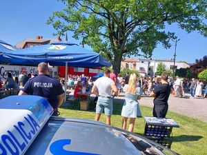 Zdjęcie w kolorze przedstawia na pierwszym planie fragment radiowozu po prawej policjant i obok niego po lewej stronie uczestnicy pikniku.