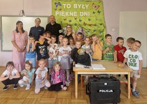 Zdjęcie w kolorze, przedstawia grupę dzieci pozujących do zdjecia. za nimi w lewym górnym rogu przedszkolanka oraz policjantka i policyjny technik. W tle zielona ścianka z napisem &quot;To były piekne dni&quot;