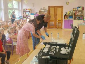 Zdjęcie w kolorze przedstawia technika policyjnego który pochylony nad stolikiem pokazuje przedszkolance jak zabezpieczyc odcisk palca.