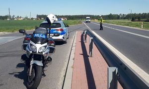 Zdjęcie w kolorze przedstawia motocykl i samochód policyjny po lewej stronie zaparkowane na zatoczce do kontroli pojazdów, w oddali policjant na jezdni daje znać sygnalizatorem do zatrzymania nadjeżdżającemu białemu busowi. W tle drzewa i łąki.
