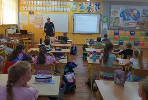 Zdjęcie w kolorze, przedstawia uczniów w szkolnych ławkach z przodu policjant.