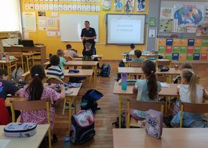 Zdjęcie w kolorze przedstawia uczniów w szkolnych ławkach, z przodu na środku klasy  stoi policjant
