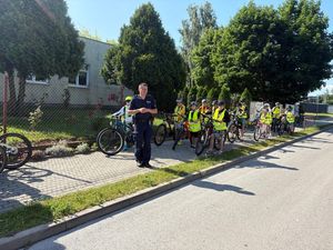 Zdjęcie w kolorze przedstawia uczniów w kaskach na głowach z rowerami stojących na chodniku przy szkole , obok stoi policjant.