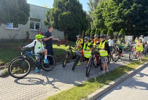 Zdjęcie w kolorze przedstawia uczniów w kaskach na głowach z rowerami stojących na chodniku przy szkole , obok stoi policjant.