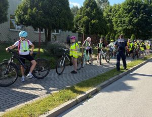 Zdjęcie w kolorze przedstawia uczniów w kaskach na głowach z rowerami stojących na chodniku przy szkole , obok stoi policjant.