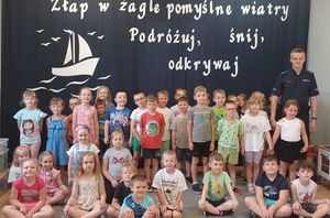 Zdjęcie w kolorze przedstawia przedszkolaków pozujących do zdjęcia na granatowym tle z napisem &quot; Złap w żagle pomyślne wiatry, podróżuj, śnij, odkrywaj&quot;. Obok dzieci stoi policjant.