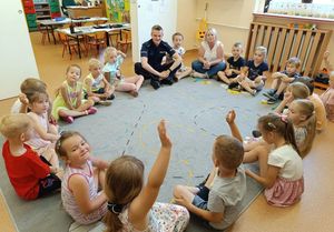 Zdjęcie w kolorze przedstawia przedszkolaków siedzących na jasnym dywanie w tak zwanym kółku, razem z nimi siedzi policjant.