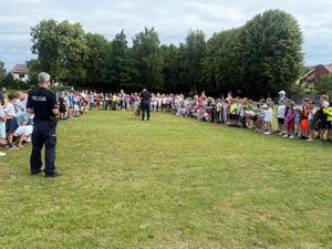 Zdjęcie w kolorze przedstawia policjanta z psem na boisku szkolnym którego otaczają uczniowie.