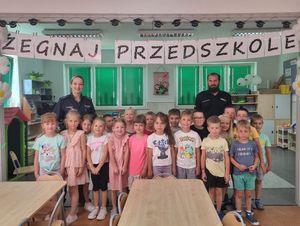 Zdjęcie w kolorze, przedstwia przedszkolaków pozujących do zdjęcia, za nimi stoi policjant i policjantka nad grupą widnieje napis &quot; Żegnaj Przedszkole&quot;