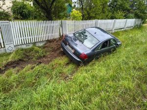 Ciemny samochód osobowy w rowie. Lewym bokiem stoi przy jasnym, betonowym ogrodzeniu posesji. Samochód lekko przychylony na prawy bok.