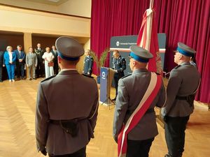 Zdjęcie w kolorze, przedstawia Komendanta Powiatowego Policji w Oleśnie podczas przemówienia w sali Miejskiego Domu Kultury w Oleśnie, z lewej strony stoi Zastępca Komendanta Wojewódzkiego insp. Krzysztof Buchała, za nimi ścianka z tkaniny z napisem Komenda Powiatowa Policji w Oleśnie. Po prawej stronie fragment Pocztu Sztandarowego oleskiej policji.
