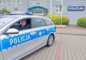 Zdjęcie w kolorze, przedstawia policjanta siedzącego w radiowozie z otwartą przednią szybą. Za pojazdem budynek Komendy Powiatowej Policji w Oleśnie.