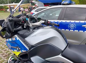 Zdjęcie w kolorze przedstawiana pierwszym planie fragment motocykla policyjnego, tuż za nim fragment samochodu policyjnego, w tle odwiedzający.