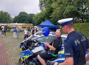 Zdjęcie w kolorze przedstawia dorosłego mężczyznę siedzącego na policyjnym motocyklu, obok stoi policjant.