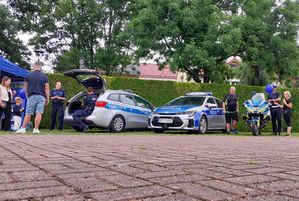 Zdjęcie w kolorze przedstawia kilka osób odwiedzających stoisko policyjne.  po prawej stoją dwa samochody i motocykl policji.