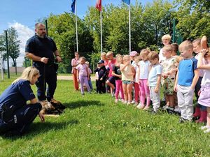 Zdjęcie w kolorze, przedstawia policjantkę i policjanta na terenie zielonym przy szkole, na przeciw grupa kilkudziesięciu dzieci.