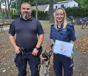 Zdjęcie w kolorze, przedstawia policjantkę i policjanta pozujących do zdjęcia, policjant trzyma na smyczy psa a policjantka w dłoniach trzyma rysunek przedstawiający psa.