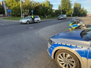 Zdjęcie w kolorze przedstawia na pierwszym planie fragment radiowozu i policyjnego motocykla, dalej na jezdni policjant kontroluje trzeźwość kierującego pojazdem osobowym.