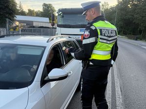 Zdjęcie w kolorze przedstawia na jezdni policjant kontroluje trzeźwość kierującego pojazdem osobowym. Tuz za nim stoi ciężarówka.
